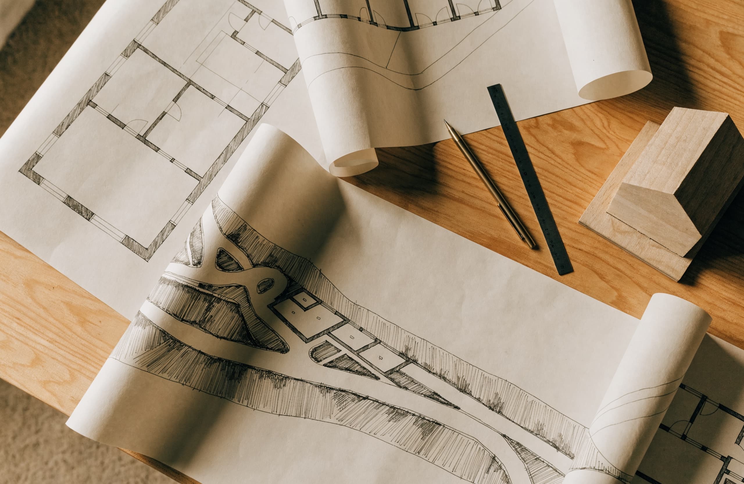Overhead flat lay of unrolled architectural floor plans and a site sketch on a timber table, with a pencil, ruler and small massing model